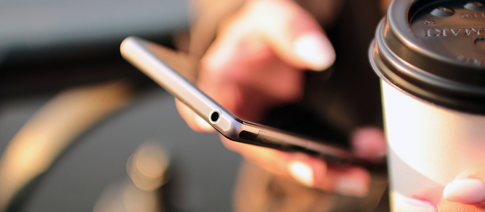 Hände halten ein Smartphone, daneben ein Kaffeebecher zum Mitnehmen, Hintergrund unscharf